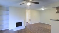 This single-story home in Bedford, Texas offers a clean, functional layout with notable design details throughout. The main living area is a welcoming space anchored by a white-painted brick fireplace, which serves as a natural focal point and adds a classic, homey character to the room. The fireplace surround and raised hearth provide a timeless aesthetic that complements the overall design.

Rich, dark wood-style flooring extends across the living area, offering both durability and visual warmth. The flooring provides a strong contrast against the light-colored walls, creating a balanced and inviting atmosphere. Neutral wall tones keep the space feeling open and versatile, well-suited to a variety of décor styles.

Overhead, a multi-blade ceiling fan with an integrated light fixture provides both air circulation and ambient lighting, enhancing everyday comfort. A secondary flush-mount light fixture is also visible, ensuring the space is well-lit throughout. A wall-mounted HVAC vent is conveniently positioned for efficient climate control.

A hallway visible from the living area suggests a straightforward, accessible floor plan, connecting the main living space to additional rooms. The textured ceiling adds subtle architectural detail, rounding out this well-maintained, move-in-ready home.