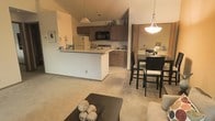 This single-floor space is designed around an open-concept layout that connects the kitchen, dining area, and living room in one cohesive flow. The kitchen features medium-toned wood cabinetry, a peninsula with counter seating, and built-in black appliances including a microwave and stovetop. The peninsula serves as both a functional workspace and an informal gathering point, separating the kitchen from the adjacent living and dining areas without closing off the space.

A pendant light hangs above the kitchen area, while a second, larger pendant fixture illuminates the dining space, where a dark-framed table and chairs provide a dedicated area for meals. The dining area transitions naturally into the living room, which is finished with neutral carpet and neutral wall tones throughout, contributing to a warm and understated ambiance.

The vaulted ceiling in the kitchen and living areas adds a sense of vertical openness, enhancing the overall feeling of spaciousness. The consistent flooring and color palette across all three zones create a unified, well-organized interior suited for comfortable, everyday living.