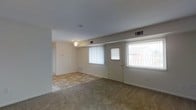 This single-story space presents an open, straightforward layout centered around a combined living and entry area. The living room is finished with wall-to-wall carpeting in a neutral beige tone, creating a warm and understated foundation. Toward the front of the space, the flooring transitions to a light-toned tile, defining a natural entryway zone adjacent to the front door. The front entry features a paneled door with a window insert, flanked by two vertically slatted window treatments that allow diffused natural light to fill the room. A ceiling fan with a light fixture is centrally positioned, providing both illumination and air circulation. HVAC vents are visible along the upper walls, indicating a ducted climate control system throughout. The walls are finished in a clean off-white tone, giving the space a neutral, move-in-ready appearance. An open passageway on the left side leads to an adjacent room, suggesting a connected and functional floor plan. Overall, the space offers a clean, practical interior with modest finishes and adequate natural light from its front-facing windows.
