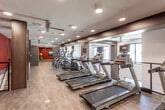 Located at 13200 133rd Ct in Overland Park, Kansas, this two-story fitness center is designed to accommodate a comprehensive range of workout activities within a well-appointed, modern environment. The facility's most striking visual element is its bold hexagonal carpet tile flooring on the lower level, featuring a dynamic pattern of red, orange, and gray tones that energizes the space and defines the strength training area.

The lower level is generously proportioned, offering soaring ceilings and large windows that invite ample natural light while providing outdoor views. This floor houses a variety of strength training machines, a dumbbell rack with open shelving storage, and interactive fitness equipment. Full-length mirrors amplify the sense of space and allow for form monitoring. Dark wood-toned columns and accents provide a sophisticated contrast to the bright flooring.

The upper level features a dedicated cardio zone lined with a row of Matrix treadmills, ellipticals, and additional equipment, all finished with smooth luxury vinyl flooring. Deep red accent walls add visual warmth, complemented by recessed lighting and ceiling fans throughout both levels. A separate studio-style room on this floor includes a wall-mounted TV, spin bike, and exercise balls, ideal for group or individual training sessions. A staircase with a modern cable railing connects both floors seamlessly.