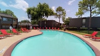 This outdoor amenity area centers around a generously sized freeform swimming pool with clear turquoise water, enclosed by a wrought iron security fence for safety. The pool deck is finished with brick-edged concrete and lined with vibrant red lounge chairs, providing ample space for sunbathing and relaxation. Mature trees surround the perimeter, offering natural shade and a pleasant, park-like atmosphere under open skies.

Adjacent to the pool is a single-story amenity building with vertical board-and-batten siding in a neutral cream tone, accented by dark brown window and door trim. The building features multiple windows along its exterior facade, allowing natural light into the interior. A covered concrete patio runs alongside the building, furnished with picnic tables and shaded by a teal umbrella, creating an inviting outdoor gathering space. A playground structure is visible in the background, adding to the recreational offerings of the area.

The overall environment is designed for community outdoor living, combining the pool, lounge area, picnic space, and play zone into a cohesive, functional amenity hub surrounded by lush greenery and open skies.
