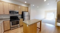 This multi-floor residence in the United States is thoughtfully designed across three levels, offering generous living space with a clean, modern aesthetic throughout. The kitchen serves as a functional centerpiece, featuring warm honey-toned wood cabinetry, stainless steel appliances including a gas range, built-in microwave, and a full-size refrigerator. A granite-topped island anchors the kitchen, providing additional prep space and casual seating. Light hardwood flooring runs through the kitchen and flows seamlessly into the adjacent open living area, creating a cohesive and airy feel. Recessed lighting throughout ensures the space is well-lit, complementing the neutral white walls that lend a bright and welcoming atmosphere. The open-concept layout between the kitchen and living area promotes easy interaction and versatile use of space. With three floors, the residence offers ample room for a variety of living arrangements, providing dedicated areas for relaxation, entertaining, and privacy. The overall design balances practicality with a contemporary style, making it a comfortable and functional home.