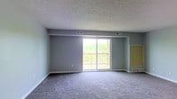 This single-floor space is laid out in a practical, open configuration that connects its key living areas in a logical flow. The floor plan reveals a generously sized main living area that serves as the centerpiece of the unit, complemented by a kitchen and several smaller rooms positioned toward one end of the layout.

The living area is carpeted throughout and finished with neutral gray-blue walls, creating a calm and understated atmosphere. A textured ceiling adds a classic residential character to the space. The standout feature of the living area is a large sliding glass door that opens directly onto a wood-railed deck. The deck overlooks an open landscape with mature trees and green scenery, allowing natural light to flood the interior while providing pleasant outdoor views.

The kitchen, visible in the floor plan model, includes cabinetry along the walls and appears compact but functional. Adjacent rooms offer additional private spaces suitable for bedrooms or storage. Multiple HVAC vents are distributed across the ceiling, indicating a forced-air climate control system throughout the unit.

Overall, the space presents a straightforward, functional design with a strong connection to the outdoors through its deck access and expansive sliding doors.