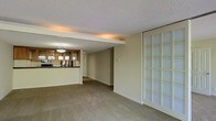 This single-floor space presents an open, connected layout that integrates the living area and kitchen into a cohesive floor plan. The carpeted living area is generously proportioned, offering a comfortable and versatile space suited for a variety of furniture arrangements. A notable architectural feature is the structural beam that visually separates the living zone from the kitchen area while maintaining an open feel throughout.

The kitchen is positioned at the rear of the space and is defined by warm wood cabinetry with glass-front upper doors, a dark countertop surface, and pendant lighting that provides focused illumination over the counter. Appliances are visible, contributing to the kitchen's functional character.

A prominent frosted glass panel door or partition along one wall introduces filtered natural light into the living area while maintaining privacy, adding a distinctive design element. The textured popcorn ceiling and cove lighting along the beam add depth and warmth to the overall ambiance. Neutral wall tones and beige carpeting throughout create a calm, understated aesthetic. A hallway visible from the main living area suggests additional rooms beyond the primary space.
