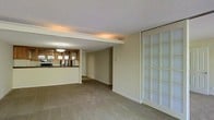 This single-floor space presents an open, connected layout that integrates the living area and kitchen into a cohesive floor plan. The carpeted living area is generously proportioned, offering a comfortable and versatile space suited for a variety of furniture arrangements. A notable architectural feature is the structural beam that visually separates the living zone from the kitchen area while maintaining an open feel throughout.

The kitchen is positioned at the rear of the space and is defined by warm wood cabinetry with glass-front upper doors, a dark countertop surface, and pendant lighting that provides focused illumination over the counter. Appliances are visible, contributing to the kitchen's functional character.

A prominent frosted glass panel door or partition along one wall introduces filtered natural light into the living area while maintaining privacy, adding a distinctive design element. The textured popcorn ceiling and cove lighting along the beam add depth and warmth to the overall ambiance. Neutral wall tones and beige carpeting throughout create a calm, understated aesthetic. A hallway visible from the main living area suggests additional rooms beyond the primary space.