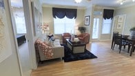 This single-story space centers around a well-appointed living area that combines classic architectural details with a warm, inviting atmosphere. Hardwood flooring extends throughout, lending a cohesive and polished look to the space. Crown molding frames the ceiling, adding an elegant finishing touch, while a ceiling fan provides both function and a subtle decorative element.

The living room is generously sized, accommodating a neutral sofa dressed with decorative throw pillows and a patterned blanket, paired with two rich brown leather accent chairs arranged around a rectangular coffee table. A dark area rug anchors the seating arrangement, creating a defined and cozy gathering space.

Multiple windows, dressed with navy blue valances over white blinds, allow ample natural light to filter into the room while maintaining privacy. A glass-paneled door on one side of the room further enhances the brightness and offers a connection to the outdoors. Wall art and decorative lamps add personality and warmth throughout. The overall ambiance is comfortable and tastefully styled, with a balance of traditional detailing and approachable design.