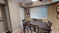 This single-floor space is thoughtfully organized across several distinct areas, each serving a specific purpose. The living room is a welcoming retreat, featuring two plush teal sofas accented with patterned throw pillows, set against cool grey walls. Vertical blinds on a large window allow for adjustable natural light, while soft carpeting underfoot adds warmth and comfort. A wall mirror enhances the sense of depth in the room.

The dining area is appointed with a rustic, dark wood table and matching chairs, offering seating for multiple guests. A ceiling fan with a light fixture provides both airflow and ambient lighting. Vertical blinds on a prominently sized window allow natural light to filter in. Framed certificates and plaques displayed on the walls lend a professional character to the space.

The kitchen is efficiently designed with warm oak cabinetry, black appliances including a gas range and refrigerator, a sink, and light wood-look flooring. An electrical panel and a water cooler are also conveniently situated here. A connecting hallway, accented with neutral tones and decorative artwork, links the spaces together, contributing to a cohesive and practical overall layout.