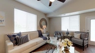 This single-floor residence offers a thoughtfully designed layout that balances comfort and functionality throughout. The living room serves as the heart of the home, featuring a vaulted tray ceiling with a ceiling fan, warm hardwood-style flooring, and multiple large windows dressed with white blinds that flood the space with natural light. The open-concept kitchen connects seamlessly to the living and dining areas, showcasing rich dark mahogany cabinetry, granite-style countertops, black appliances, and a breakfast bar with seating — ideal for casual dining or entertaining. The space accommodates multiple bedrooms, each carpeted for comfort and fitted with windows that bring in ample daylight. The bedrooms offer generous proportions, providing space for full furnishings and personal touches. The bathroom is well-equipped with a dark wood vanity, granite countertop, a large mirror, and wood-style flooring, complemented by a shower-tub combination. Neutral warm tones carry throughout the residence, creating a cohesive and inviting ambiance. The overall design reflects a practical yet attractive aesthetic, with consistent finishes and architectural details that lend the space a polished, residential character.