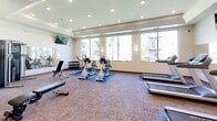 Located at 2915 Bluestem Dr in West Fargo, North Dakota, this fitness center is a well-appointed, single-floor facility designed to support a comprehensive workout experience. The space is bright and open, benefiting from large windows that allow abundant natural light to fill the room while offering views of the surrounding building exterior. Recessed lighting throughout the tray ceilings further ensures the space is well-lit at all times.

The cardio area is a standout feature, equipped with multiple treadmills and elliptical machines positioned in rows, each offering ample space for comfortable use. Wall-mounted flat-screen televisions are strategically placed throughout the room, providing entertainment during workouts. Full-length mirrors line one wall, enhancing the sense of space and allowing users to monitor their form.

The strength and flexibility zone includes a dumbbell rack stocked with a range of weights, adjustable benches for free weight exercises, and stationary cycling bikes for low-impact cardio training. A dedicated accessory area houses stability balls in various sizes, a foldable workout mat, foam rollers, and resistance tools. A water cooler station is conveniently available to keep users hydrated. The carpet flooring with blue mat overlays adds comfort underfoot throughout the entire facility.