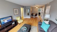This well-appointed single-floor residence offers a thoughtfully designed layout that seamlessly connects the living, dining, and entry areas into one cohesive and inviting space. Rich hardwood flooring flows throughout, lending warmth and continuity to every room.

The living area is comfortably sized, featuring neutral gray walls that provide a versatile backdrop for furnishings. A ceiling fan overhead ensures year-round comfort. A full-length mirror and tasteful wall art add character, while the open sightlines create a sense of spaciousness that extends naturally into the dining area.

The dining space is positioned centrally within the home, accommodating a round table and seating for small gatherings. Its placement between the living area and the rest of the home makes it an ideal hub for everyday use and entertaining alike.

Multiple doorways lead to additional rooms and hallways, suggesting a practical and well-organized floor plan. The entryway is clearly defined, contributing to the overall sense of flow and structure throughout the home.

Natural light enhances the bright, airy ambiance, complementing the warm tones of the hardwood floors and the cool gray palette of the walls. The overall aesthetic is clean, modern, and welcoming — a space that balances style with everyday functionality.