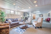 This single-floor community space offers a well-appointed layout divided into several functional zones. The lounge area is warmly furnished with a plush sofa, floral accent chairs, and a round glass-topped coffee table set atop a decorative area rug, creating an inviting atmosphere for relaxed gathering. A fireplace anchors one end of the lounge, complemented by a console and decorative touches including a large wall mirror and indoor greenery. Natural light filters in through a row of horizontal windows running along the paneled walls, giving the space a bright, airy feel despite its below-grade setting. The paneled walls, finished in a soft cream tone, lend a classic, cottage-like character throughout. A high-top table with bar stools provides a casual seating option near the center of the space, ideal for conversation or light activity. A dedicated recreation room features a full-size ping-pong table surrounded by red upholstered chairs, offering ample space for games and events. A small kitchenette area with wooden cabinetry, shelving, and a countertop workspace rounds out the utility of the space. Recessed and flush-mount lighting throughout ensures consistent illumination across all areas.