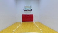 This indoor basketball court offers a dedicated athletic space designed for competitive and recreational play. The court features a classic hardwood-style floor finished in a warm natural tone, complete with clearly marked white boundary and key lines that define the playing area with precision.

At the focal end of the court, a wall-mounted basketball hoop with a transparent glass backboard and red accents provides a regulation-style shooting target. Directly beneath the hoop, a set of bold red padded wall panels runs the width of the end wall, offering an important safety buffer for players driving to the basket.

The surrounding walls are finished in clean white, contributing to a bright and open atmosphere. The single-level layout keeps the space straightforward and functional, with the high ceiling providing ample vertical clearance for play. The overall design prioritizes athletic utility, making this court well-suited for organized games, skill training, or casual recreational use. The combination of safety padding, quality flooring, and proper court markings reflects a thoughtfully equipped indoor sports facility.