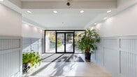 This single-floor lobby presents a clean, modern aesthetic defined by its spacious, open layout and carefully considered design details. Large-format light beige ceramic tiles cover the floor throughout, lending a sense of continuity and generous scale to the space. The walls feature distinctive grey beadboard wainscoting that runs the full length of the lobby, adding a refined architectural character while maintaining a neutral, welcoming palette. Above the wainscoting, bright white walls and a white ceiling with recessed pot lighting create a well-lit, airy atmosphere.

At the front of the lobby, full-height glass entry doors framed in black allow natural sunlight to pour in, casting soft shadows across the floor and offering a clear view to the landscaped exterior. Potted tropical plants positioned near the entrance introduce a touch of greenery and warmth to the otherwise minimal interior.

Moving deeper into the lobby, a raised platform leads to a centrally positioned elevator finished in light wood and brushed stainless steel, flanked by grey utility doors. A security camera and smoke detector are mounted at the ceiling, reflecting the practical, functional nature of the space. Overall, the lobby exudes a tidy, professional ambiance suited to a well-maintained residential or commercial building.