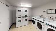 This shared laundry room is a functional, single-floor facility designed to efficiently serve multiple users. The space features a combination of stacked and front-loading commercial-grade washing machines and dryers, all finished in white and equipped with digital controls and coin-operated payment systems. The machines are numbered for easy identification, and informational signage is posted on the walls to guide users through proper operation and payment methods.

A built-in laminate folding counter runs along one wall, providing a convenient surface for sorting and folding laundry. A corkboard bulletin board is mounted nearby, offering a place for notices and instructions. The room is finished with light-colored vinyl flooring, white-painted concrete block walls, and standard ceiling-mounted fluorescent lighting that ensures adequate illumination throughout the space.

Two solid-core doors with automatic closers provide entry and exit points, maintaining privacy and security. The layout is straightforward and efficient, maximizing usable floor space while keeping all machines accessible. Overall, the room is clean, well-organized, and purpose-built to meet the practical laundry needs of its users.