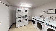 This shared laundry room is a functional, single-floor facility designed to efficiently serve multiple users. The space features a combination of stacked and front-loading commercial-grade washing machines and dryers, all finished in white and equipped with digital controls and coin-operated payment systems. The machines are numbered for easy identification, and informational signage is posted on the walls to guide users through proper operation and payment methods.

A built-in laminate folding counter runs along one wall, providing a convenient surface for sorting and folding laundry. A corkboard bulletin board is mounted nearby, offering a place for notices and instructions. The room is finished with light-colored vinyl flooring, white-painted concrete block walls, and standard ceiling-mounted fluorescent lighting that ensures adequate illumination throughout the space.

Two solid-core doors with automatic closers provide entry and exit points, maintaining privacy and security. The layout is straightforward and efficient, maximizing usable floor space while keeping all machines accessible. Overall, the room is clean, well-organized, and purpose-built to meet the practical laundry needs of its users.
