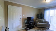 This single-floor space features a living area finished in warm, neutral tones that create an inviting atmosphere. The walls are painted in a soft tan hue, complemented by clean white crown molding that adds a touch of classic detail along the ceiling perimeter. The ceiling itself features a textured popcorn finish, a characteristic element of the space.

Light-toned tile flooring runs throughout, offering durability and easy maintenance, while a patterned area rug introduces a subtle decorative layer to the living zone. The layout accommodates essential furnishings comfortably, including seating and an entertainment setup supported by a dark wood and glass media stand.

A six-panel white entry door with a standard lockset serves as the main point of entry, positioned along one wall of the living area. A lush indoor plant in the corner adds a natural, organic element to the space, enhancing the overall warmth of the room. The combination of neutral wall color, classic trim work, and practical flooring gives the space a tidy, functional character well-suited for day-to-day living.