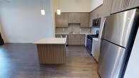 This single-floor space showcases a thoughtfully designed open-concept layout, centered around a modern kitchen that serves as the heart of the home. The kitchen is outfitted with a full suite of stainless steel appliances, including a gas range, microwave, dishwasher, and refrigerator, all complemented by sleek, warm-toned flat-front cabinetry that extends to the upper walls, providing generous storage. A hexagonal tile backsplash adds a refined decorative touch, while light granite countertops offer a bright contrast to the rich, dark hardwood-style flooring that runs throughout the space. A central kitchen island with a matching wood-panel base and stone countertop provides additional workspace and casual seating potential. Pendant lighting hangs above the island, casting a warm, focused glow that enhances the contemporary ambiance. The soaring ceiling height lends an airy, open feel to the kitchen and adjacent living area, contributing to the overall sense of spaciousness. The neutral color palette throughout creates a clean, modern aesthetic that is both inviting and functional, making this a well-appointed space suited for everyday living and entertaining alike.