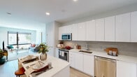 This contemporary single-floor residence showcases a thoughtfully designed open-concept layout that seamlessly connects the kitchen, dining, and living areas. The kitchen is a standout, featuring high-gloss white cabinetry extending to the ceiling, a light quartz countertop island, a neutral subway tile backsplash, and a full suite of stainless steel appliances including a range, microwave, dishwasher, and refrigerator. Light wood-tone flooring runs throughout, lending warmth and visual continuity to the space.

The living area is bathed in natural light through expansive floor-to-ceiling windows with views of the surrounding neighborhood, creating an airy, connected atmosphere. A low-profile media console anchors the room, complemented by a comfortable seating arrangement beneath the oversized windows.

The bedrooms are serene and well-proportioned, each finished with soft neutral walls and light hardwood flooring. One bedroom benefits from a large window that frames an outdoor view, while the other features a round wall mirror that enhances its sense of space.

The bathroom is clean and modern, highlighted by a glass-enclosed walk-in shower finished with crisp white subway tile and polished chrome fixtures, reflecting the home's consistent contemporary aesthetic throughout.
