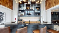 Located at 141 Stobhill Ln in Fuquay-Varina, NC, this thoughtfully designed amenity space offers a compelling blend of modern aesthetics and functional versatility across a single expansive floor. The entrance opens into a dramatic common area distinguished by soaring vaulted ceilings, polished concrete floors, and an abundance of natural light streaming through large windows. The kitchen and lounge area serves as the social centerpiece, anchored by a sleek dark island with leather bar seating, warm wood cabinetry, a white tile backsplash, and a dramatic floor-to-ceiling open shelving unit with integrated TV and pendant globe lighting. An accent wall clad in natural wood paneling adds warmth and texture to the open-plan space. The living area features a statement fireplace with a mounted TV, flanked by built-in shelving, a plush sectional sofa, a shuffleboard table, and striking sculptural pendant lights. Intimate booth-style seating with bold patterned wallpaper provides cozy nooks for smaller gatherings. A dining area with high-top tables and billiard-themed artwork connects seamlessly to an outdoor patio through full-glass French doors. The dedicated fitness center is outfitted with professional-grade cardio equipment, a mounted TV, a water station, and motivational wall art, offering a bright, well-organized workout environment.