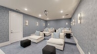 Located in Winter Garden, FL, this dedicated home theater room is a thoughtfully designed entertainment space that delivers a cinematic experience in a refined residential setting. The room is anchored by a large projection screen mounted on one wall, paired with a ceiling-mounted projector, creating an immersive viewing setup. The seating area features multiple oversized cream-colored modular lounge chairs and chaises arranged in rows, all dressed with coordinating blue and white accent pillows, ensuring optimal comfort for every viewer. Dark charcoal ottomans are positioned throughout for additional flexibility. The walls are finished in a soft blue-toned geometric patterned wallcovering, lending a sophisticated, theater-inspired ambiance. Elegant vertical sconce lighting is evenly distributed across all walls, providing a warm, dimmable glow that enhances the cinematic atmosphere. A dedicated snack and beverage station is built into one corner, featuring wood cabinetry, a beverage refrigerator, a microwave, and pendant lighting above — all set against a bold hexagonal tile backsplash accent wall. The flooring transitions from large-format white tile near the entry to plush gray carpet in the main seating area. A glass-paneled door with a "Reserved" sign adds a charming, venue-inspired touch to this well-appointed private theater.