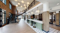This expansive communal lobby showcases a masterful blend of industrial heritage and modern design. Soaring double-height ceilings anchor the space, drawing the eye upward to a mezzanine level framed by a sleek black metal railing and a dramatic open-tread staircase. Exposed brick walls run throughout, adding warmth and texture that contrasts beautifully with the crisp white upper walls and ceilings.

At the heart of the space is a long, sage-green bar counter topped with white quartz, accompanied by black and gold barstools and framed by a distinctive black-and-white hexagonal tile floor. Geometric pendant lanterns and a cascade of globe pendants overhead create a layered, inviting lighting scheme. Natural light pours in generously through oversized black-framed windows, offering views of the surrounding greenery.

The open floor plan accommodates distinct seating vignettes, including a lounge area with stylish accent chairs and a casual dining zone with industrial-style metal furniture. Wood-paneled millwork detailing adds a refined, crafted touch near the service area, where sage-green lower cabinetry with stainless steel appliances continues the cohesive color palette. The overall ambiance is sophisticated yet approachable, balancing historic character with contemporary comfort.