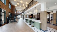 This expansive communal lobby showcases a masterful blend of industrial heritage and modern design. Soaring double-height ceilings anchor the space, drawing the eye upward to a mezzanine level framed by a sleek black metal railing and a dramatic open-tread staircase. Exposed brick walls run throughout, adding warmth and texture that contrasts beautifully with the crisp white upper walls and ceilings.

At the heart of the space is a long, sage-green bar counter topped with white quartz, accompanied by black and gold barstools and framed by a distinctive black-and-white hexagonal tile floor. Geometric pendant lanterns and a cascade of globe pendants overhead create a layered, inviting lighting scheme. Natural light pours in generously through oversized black-framed windows, offering views of the surrounding greenery.

The open floor plan accommodates distinct seating vignettes, including a lounge area with stylish accent chairs and a casual dining zone with industrial-style metal furniture. Wood-paneled millwork detailing adds a refined, crafted touch near the service area, where sage-green lower cabinetry with stainless steel appliances continues the cohesive color palette. The overall ambiance is sophisticated yet approachable, balancing historic character with contemporary comfort.