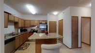 This single-floor space centers around a well-appointed kitchen designed for both practicality and comfort. The kitchen features warm honey-toned wood cabinetry that extends along the perimeter walls, offering generous storage. Stainless steel appliances, including a refrigerator and electric range with overhead ventilation, provide a modern contrast to the traditional cabinetry. A central island with a granite countertop serves as a natural gathering point and casual dining area, complemented by seating on one side. The granite surfaces display neutral tones with subtle veining, adding a touch of elegance to the space. Light hardwood-style flooring runs throughout, contributing to a bright, cohesive aesthetic. Flush-mounted ceiling light fixtures provide consistent, even illumination across the kitchen area. The layout flows openly toward adjacent doorways, suggesting connectivity to other areas of the unit. The overall ambiance is warm and inviting, blending functional design elements with classic finishes that create a comfortable, everyday living environment.