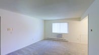 This single-floor space presents a clean, minimalist layout centered around an open living area. The room features neutral beige walls and a textured popcorn ceiling, contributing to a simple, understated aesthetic. Wall-to-wall carpet in a light gray tone covers the floor, providing a soft, comfortable surface underfoot and adding warmth to the overall atmosphere.

Natural light enters the space through a centrally positioned window fitted with vertical blinds, allowing for adjustable light control throughout the day. Positioned beneath the window is a wall-mounted air conditioning unit, offering a dedicated cooling solution for the room. Along the base of the wall, a baseboard heater runs the length of the exterior wall, providing an efficient heating source during colder months. Together, these features ensure year-round climate comfort.

The walls are smooth and unadorned, offering a blank canvas for personalization. The open layout maximizes the usable floor area, providing flexibility in how the space can be arranged and utilized. Overall, the room conveys a functional, straightforward design well-suited for a variety of living arrangements.