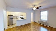 This single-floor space offers a bright and functional open-concept layout that seamlessly connects the kitchen and living area. The living space is grounded by classic parquet hardwood flooring, which adds warmth and character throughout. A ceiling fan with an integrated light fixture is centrally mounted, enhancing airflow and providing ambient lighting.

The kitchen is thoughtfully designed with white cabinetry that extends to the ceiling, offering generous storage. A granite-topped peninsula counter serves as a natural partition between the kitchen and living area, ideal for casual dining or food preparation. The kitchen is equipped with a stainless steel sink, a gas range, and an over-the-range ventilation unit.

Natural light enters the living area through a double-hung window, which overlooks greenery outside. A classic cast-iron radiator sits beneath the window, providing efficient heating. Crown molding trims the ceiling, lending a refined architectural detail to the space. A doorway visible from the main area hints at additional rooms beyond, suggesting a practical and well-connected floor plan. The overall ambiance is clean, bright, and move-in ready, with timeless finishes that offer a welcoming residential feel.