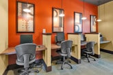 Located at 34875 Pourroy Rd in Winchester, CA, this single-room business center office is a thoughtfully designed workspace that balances functionality with stylish aesthetics. The room features a bold two-tone color scheme, with rich terracotta-orange accent walls contrasting against softer cream and sage green surfaces, creating a warm yet professional atmosphere. Dark wood baseboards and trim add a polished finishing touch throughout.

The office is equipped with multiple built-in workstation cubicles along the walls, each featuring a desktop computer, keyboard, and ergonomic task chair — ideal for focused individual work. A multifunction printer station is also available, making the space fully equipped for everyday office needs. Pendant lighting suspended from a dark ceiling provides warm, directed illumination over the workstations, complemented by a recessed ceiling light and natural light streaming in through a window fitted with blinds.

The space is further enhanced by framed contemporary artwork on the terracotta walls and a striking large-scale metal branch wall sculpture, adding an artistic dimension to the room. Light-toned tile flooring grounds the interior, while a glass-paneled entry door with a sidelight connects the office to the broader facility. A bulletin board and intercom panel round out the practical features of this efficiently arranged, characterful office space.