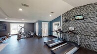 This fitness center is a thoughtfully designed, single-level facility offering a comprehensive workout environment across multiple interconnected zones. The space is painted in cool teal and gray tones, complemented by dark rubber flooring and a textured ceiling with recessed lighting throughout.

The cardio area is a standout feature, housing multiple True-brand treadmills, elliptical machines, and a stationary bike positioned along a striking mosaic tile accent wall. Mounted flat-screen televisions above the equipment provide entertainment during workouts. Natural light pours in through several windows and glass-panel doors that open toward an outdoor pool area, creating a bright, inviting atmosphere.

The strength training section is well-appointed with Precor cable machines, a multi-station weight system, a dumbbell rack, adjustable benches, and additional functional training tools including a punching bag, kettlebells, and balance equipment. A dedicated open floor space supports functional fitness activities.

A compact kitchenette serves the facility, featuring warm wood cabinetry, granite countertops, a double stainless steel sink, and a matching mosaic tile backsplash — offering convenience for post-workout refreshment. Overall, the fitness center delivers a professional and energizing environment suited for a wide range of training routines.