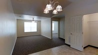 This single-floor residence offers a straightforward and functional layout designed for comfortable everyday living. The main living area is finished with plush wall-to-wall carpeting, creating a warm and cozy atmosphere, while the entryway features light-toned tile flooring that provides a clean transition into the home. An elegant multi-arm chandelier serves as a central focal point overhead, providing ample illumination to the space, while a ceiling fan in the adjoining area ensures year-round comfort and air circulation. Natural light filters in through a large window fitted with blinds, brightening the living space and giving it an airy, open feel. Neutral-toned walls throughout contribute to a calm and versatile aesthetic that accommodates a variety of décor styles. A hallway visible from the main living area leads further into the home, connecting additional rooms through clean white-trimmed doorways. Flush baseboards and wall vents reflect the practical construction of the space. The overall ambiance is clean, understated, and move-in ready, making it a solid and dependable living space suited for a range of occupants.