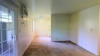 This single-story space features an open, straightforward layout with a living area that flows into an adjoining room, likely a kitchen or dining area, visible through an open passage in the far wall. The living area is finished with wall-to-wall carpeting in a neutral beige tone, while the transition to the adjacent space features a darker flooring. Walls are painted in a light, muted green tone, contributing to a calm, understated ambiance throughout. A wall-mounted air conditioning unit is installed on the left wall, providing climate control, and a window nearby allows for natural light to filter into the space. A ceiling fan with an integrated light fixture is positioned in the adjoining area, offering both airflow and overhead illumination. The ceilings are flat and painted white, maintaining a clean, simple aesthetic. The overall layout is compact yet functional, with the open passage between rooms promoting a sense of connectivity. The space appears unfurnished, allowing a clear view of its structural elements and offering flexibility for various furniture arrangements and uses.