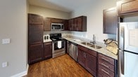 This single-floor unit presents a cohesive, neutral design palette carried consistently throughout its living spaces. The open-concept layout connects the living area to the kitchen, where rich, dark-stained cabinetry provides striking contrast against light granite countertops. Stainless steel appliances—including a refrigerator, range, microwave, and dishwasher—complement the wood-look flooring that runs through the kitchen and adjoining areas. A double-basin stainless steel sink with a modern faucet adds practical functionality to the kitchen workspace.

The living and bedroom areas feature plush, neutral-toned carpeting underfoot, contributing to a warm and comfortable atmosphere. Walls throughout are finished in a soft greige tone, complemented by crisp white baseboards and trim. A textured ceiling adds subtle visual interest overhead, while multiple electrical outlets are conveniently distributed throughout.

Natural light enters through window blinds, and a 3D floor plan view reveals a thoughtfully segmented layout with multiple rooms connected by interior doorways. The bathroom is finished with wood-look flooring, a tub-shower combination with a chrome curtain rod, a granite-topped vanity with dark cabinetry, and clean white fixtures, maintaining the consistent aesthetic found throughout the unit.