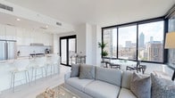 This contemporary high-rise residence is defined by clean lines, a crisp white palette, and floor-to-ceiling windows that frame expansive city skyline views. The open-concept living and dining area is bright and airy, with light hardwood flooring flowing seamlessly throughout. Large black-framed windows flood the space with natural light and provide stunning urban panoramas from multiple vantage points, complemented by access to a private balcony.

The sleek, modern kitchen features high-gloss white cabinetry, a large island with bar seating, stainless steel appliances, and a subway tile backsplash — ideal for both everyday living and entertaining. The kitchen opens directly to the living and dining areas, reinforcing the home's open and connected layout.

The primary bedroom offers direct balcony access with sweeping views, plush carpeting, and dual closets. Its en-suite bathroom is finished in large-format light tiles and features a long floating vanity and a glass-enclosed walk-in shower. A second bedroom provides a comfortable and well-appointed retreat, served by a full bathroom with a soaking tub-shower combination and clean white finishes. Throughout, dark-framed doors add a modern contrast to the bright, neutral interiors, creating a polished and cohesive aesthetic.