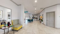 This single-floor clubhouse facility is designed with a contemporary aesthetic, defined by bold teal accent walls, crisp white surfaces, and large-format marble-look porcelain tile flooring that runs consistently throughout. The main lounge area commands attention with soaring double-height ceilings, a coffered teal-trimmed tray detail, and an oversized ceiling fan that enhances the airy atmosphere. Floor-to-ceiling windows flood the space with natural light while offering views of surrounding greenery. A striking tile-clad fireplace serves as the focal centerpiece, flanked by built-in shelving niches with yellow upholstered seating, adding vivid pops of color. Comfortable modern furnishings, including swivel chairs, sofas, and a glass coffee table, create inviting conversation areas. An adjacent dining zone features teal velvet chairs around a wood-grain table, complemented by mirrored wall accents and decorative vignettes. The fully equipped kitchen includes stainless steel appliances, light wood cabinetry, a tile backsplash, granite countertops, and pendant lighting. A dedicated private office provides a functional workspace with natural light, a full desk setup, and curated artwork. The entry corridor offers additional casual seating and connects seamlessly to all areas of the facility.