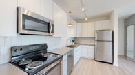 Located at 530 W Town St in Columbus, Ohio, this single-floor residence offers a thoughtfully designed open layout with clean, modern finishes throughout. The kitchen is a standout feature, outfitted with floor-to-ceiling white cabinetry, a white subway tile backsplash, quartz-style countertops, and a full suite of stainless steel appliances including a refrigerator, electric range, built-in microwave, and dishwasher. A distinctive orange pendant light and track lighting overhead add a warm, contemporary accent to the bright, functional workspace. Light hardwood flooring flows seamlessly from the kitchen into the open living area, which is bathed in natural light from a large sliding glass door that opens to the outdoors. The living space is generously proportioned with a neutral color palette of crisp whites and soft grays, lending a calm, airy ambiance. The bathroom is finished with wood-look tile flooring, a white vanity with a dark countertop, and a walk-in shower adorned with elongated vertical gray tiles that extend to the ceiling, creating a striking focal point. Throughout the unit, the design emphasizes clean lines, quality materials, and a modern aesthetic that suits a variety of lifestyles.