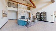 Located at 14211 Akron Ave in Rosemount, MN, this single-floor lobby lounge is a thoughtfully designed communal space that makes a bold architectural statement. The room is anchored by a dramatic floor-to-ceiling natural stone fireplace that draws the eye upward toward soaring vaulted ceilings adorned with prominent exposed wood beams. An upper-level mezzanine overlooks the main floor, adding vertical depth and a sense of grandeur to the space. A contemporary chandelier with exposed bulbs hangs gracefully above the central seating area, providing warm ambient lighting. Large-format tile flooring in a neutral gray tone spans the entire floor, lending a clean, modern foundation to the design. The entry features a full glass storefront facade, flooding the foyer with natural light and offering views to the exterior. The living area is furnished with a curved teal sofa, soft sage armchairs, and a light wood coffee table arranged over a richly patterned area rug, creating an inviting and stylish gathering zone. Two arched black metal display cabinets with glass panels serve as decorative focal points along one wall, while a built-in mailbox alcove reflects the building's functional amenities. Black-framed interior glass doors provide transitions to adjacent spaces while maintaining visual openness throughout.