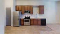 This single-floor unit offers a practical and comfortable layout designed for everyday living. The kitchen is a standout feature, outfitted with warm-toned wooden cabinetry extending to the upper walls for generous storage. Stainless steel appliances — including a refrigerator, electric range, and built-in microwave — complement the light countertops, while a black dishwasher and a modern pull-down faucet over the sink complete the functional setup. Wood-look flooring runs through the kitchen area, transitioning seamlessly into plush wall-to-wall carpeting in the adjoining living space. The living area is bright and open, benefiting from natural light that filters through a set of wide, blinds-fitted windows. Neutral wall tones and clean lines give the space a calm, understated character. The bathroom is well-appointed, featuring a soaking bathtub with a curved shower rod and built-in corner shelving, paired with a rich dark-wood vanity topped with a white countertop and a large wall mirror framed by overhead lighting. Tile flooring and crisp white walls round out the clean, functional aesthetic of the bathroom. Overall, the unit presents a cohesive, move-in-ready environment with thoughtful finishes throughout.
