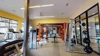 This single-floor fitness center is thoughtfully designed to accommodate a wide range of workout styles and training needs. The space is organized into several distinct zones, each tailored to a specific type of exercise. The main strength and functional training area features bold orange Octagon rig structures equipped with cable attachments, wall-mounted storage for weights, adjustable benches, and a punching bag, all set atop durable speckled rubber flooring. Linear recessed lighting and ceiling fans run the length of this section, maintaining a clean, energetic atmosphere against warm yellow walls.

The cardio zone transitions naturally from the strength area and houses a row of treadmills and elliptical machines positioned alongside floor-to-ceiling glass windows, offering views of an outdoor courtyard and filling the space with natural light. Additional resistance machines and cable units are distributed throughout this open area.

A dedicated studio room features hardwood flooring, full-wall mirrored panels framed in rich wood trim, a mounted barre, a flat-screen television, and storage for plyometric boxes and step platforms — suited for group classes, barre, and functional training. A bulletin board and check-in terminal near the entrance provide informational and access amenities for users.