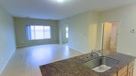 This single-floor residence offers a clean, open-concept layout designed to maximize both space and functionality. The kitchen serves as a central hub, featuring rich dark granite countertops with a stainless steel sink, providing a striking contrast against the light-toned walls and flooring throughout. The kitchen opens seamlessly into a generous living area, creating a connected and airy atmosphere ideal for everyday living and entertaining. Large windows along the exterior wall flood the living space with abundant natural light, enhancing the overall sense of openness. The smooth, light-colored hard flooring extends throughout, contributing to a cohesive and easy-to-maintain interior. Neutral wall tones in soft beige provide a versatile backdrop that complements a variety of furnishing styles. Additional rooms are accessible through interior doorways visible from the main living area, suggesting a practical and well-organized floor plan. The space presents a modern, move-in-ready aesthetic with clean lines, minimal ornamentation, and thoughtful design choices that balance comfort with contemporary style.
