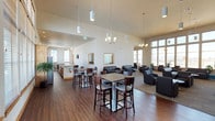This single-floor community clubhouse is designed to provide a welcoming and versatile gathering space with a polished, contemporary aesthetic. The open-concept layout seamlessly connects multiple functional zones, all unified by soaring ceilings and rich hardwood flooring throughout the main areas.

The fully equipped kitchen serves as a central hub, featuring warm knotty wood cabinetry, a granite-topped island with bar seating, stainless steel appliances including a side-by-side refrigerator and microwave, and a tasteful mosaic tile backsplash. Floor-to-ceiling windows flood the kitchen and adjacent dining area with natural light and offer views of an outdoor pool.

The dining zone accommodates both high-top and standard-height tables with ample seating, making it ideal for casual gatherings or organized events. The lounge area transitions smoothly from the dining space, featuring dark leather sofas and armchairs arranged around a striking stacked-stone fireplace with a wall-mounted flat-screen TV — a natural focal point for the room.

Additional seating nooks are positioned along the expansive window walls, inviting residents to relax in natural light. The space also includes a billiards table, adding a recreational element. Neutral tones, warm wood accents, elegant pendant and chandelier lighting, and abundant greenery contribute to an inviting, upscale ambiance throughout.