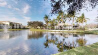 This charming single-story property is nestled within a picturesque tropical setting, offering a peaceful and inviting atmosphere that immediately captures attention. The structure sits gracefully along the edge of a calm, reflective waterway, providing stunning lakefront views that serve as a natural backdrop to the entire space. Towering palm trees and mature oak canopies surround the property, casting dappled shade and lending a lush, verdant quality to the landscape. The exterior architecture features a low-profile roofline with an open, approachable design that blends harmoniously with the natural surroundings. A covered outdoor area, likely a patio or pergola, extends from the main structure and opens toward the water, creating an ideal space for relaxation or social gatherings while taking in the scenic views. The well-manicured lawn stretches to the water's edge, enhancing the sense of openness and connection to the outdoors. The overall ambiance is one of tropical tranquility — a setting that evokes the relaxed elegance of Florida living, where natural beauty, warm sunlight, and waterfront serenity come together seamlessly to define the character of this distinctive property.