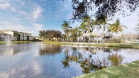 This charming single-story property is nestled within a picturesque tropical setting, offering a peaceful and inviting atmosphere that immediately captures attention. The structure sits gracefully along the edge of a calm, reflective waterway, providing stunning lakefront views that serve as a natural backdrop to the entire space. Towering palm trees and mature oak canopies surround the property, casting dappled shade and lending a lush, verdant quality to the landscape. The exterior architecture features a low-profile roofline with an open, approachable design that blends harmoniously with the natural surroundings. A covered outdoor area, likely a patio or pergola, extends from the main structure and opens toward the water, creating an ideal space for relaxation or social gatherings while taking in the scenic views. The well-manicured lawn stretches to the water's edge, enhancing the sense of openness and connection to the outdoors. The overall ambiance is one of tropical tranquility — a setting that evokes the relaxed elegance of Florida living, where natural beauty, warm sunlight, and waterfront serenity come together seamlessly to define the character of this distinctive property.