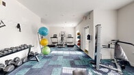Located at 6229 Thomaston Rd in Macon, Georgia, this single-floor fitness center is a well-equipped community gym designed to support a well-rounded workout routine. The space is laid out across an open, L-shaped floor plan covered in a distinctive teal and navy patterned carpet that adds a modern aesthetic to the bright, white-walled interior.

The cardio zone is anchored by multiple treadmills and an elliptical trainer, positioned beneath recessed ceiling lights that keep the area well-lit throughout the day. Natural light streams in through windows fitted with shuttered blinds, creating an airy and energizing atmosphere. A large wall-mounted mirror in one section visually expands the space and allows users to monitor their form during workouts.

The strength training area includes a multi-function cable machine, a weight bench, and a rack of dumbbells, providing options for both free-weight and machine-based resistance training. A dedicated rack holds an assortment of stability and medicine balls of varying sizes, supporting functional fitness and core training. A cable resistance attachment station rounds out the strength offerings.

Additional amenities include a wall-mounted water fountain and bottle-filling station, posted fitness center rules, and a security camera, ensuring a safe and convenient environment. The space is accessible via a French-door entrance and is finished with clean white walls and neutral trim throughout.
