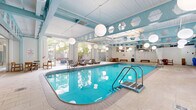 Located at 1 Franklin Town Blvd in Philadelphia, PA, this indoor recreational facility spans a generously sized single floor and centers around a large, striking indoor swimming pool with vivid turquoise water. The space features a distinctive architectural design, with bold teal structural beams, a partially skylighted ceiling that floods the pool area with natural light, and white-painted brick walls that contribute to the bright, clean aesthetic throughout.

Suspended globe pendant lights are positioned throughout the space, providing warm, even illumination that complements the natural light from the skylights and large floor-to-ceiling glass doors and windows. These glass panels open to an outdoor area, offering views of greenery and maintaining a connection to the exterior environment.

The pool deck is finished in neutral, light-toned tile flooring and is furnished with cushioned lounge chairs and benches for relaxation. Seating areas with round tables and wood-framed chairs are available poolside, creating comfortable spaces for gathering. An adjacent fitness room, visible through interior glass partitions, provides additional recreational options. The overall ambiance is bright, clean, and functional, making this a well-appointed amenity space designed for active recreation and leisure.