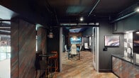 This single-floor office space blends industrial and contemporary design into a cohesive, sophisticated work environment. The exposed ceiling with visible ductwork and dark painted finishes establishes an urban, loft-inspired aesthetic carried throughout the space. Multicolored, textured carpet tiles in warm rust, amber, and grey tones anchor the floor and provide a striking visual contrast against the predominantly dark walls and ceiling.

The conference room serves as a focal point, enclosed by glass partitions that allow natural light to filter through while maintaining a sense of privacy. Inside, a long dark conference table is surrounded by sleek white high-back chairs, and a large wall-mounted display screen dominates one end of the room. A sculptural, oversized pendant light fixture adds a dramatic design element overhead, complementing a recessed panel of richly oxidized, multicolored accent material on the ceiling.

Adjacent to the conference room, a casual collaboration zone features marble-topped bistro tables with bold orange bar stools atop brass-finished bases. A lounge nook with a high-backed upholstered booth seat and a beverage station with a display cabinet and under-counter refrigeration provide additional function. The overall ambiance is intentional, moody, and design-forward.