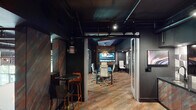 This single-floor office space blends industrial and contemporary design into a cohesive, sophisticated work environment. The exposed ceiling with visible ductwork and dark painted finishes establishes an urban, loft-inspired aesthetic carried throughout the space. Multicolored, textured carpet tiles in warm rust, amber, and grey tones anchor the floor and provide a striking visual contrast against the predominantly dark walls and ceiling.

The conference room serves as a focal point, enclosed by glass partitions that allow natural light to filter through while maintaining a sense of privacy. Inside, a long dark conference table is surrounded by sleek white high-back chairs, and a large wall-mounted display screen dominates one end of the room. A sculptural, oversized pendant light fixture adds a dramatic design element overhead, complementing a recessed panel of richly oxidized, multicolored accent material on the ceiling.

Adjacent to the conference room, a casual collaboration zone features marble-topped bistro tables with bold orange bar stools atop brass-finished bases. A lounge nook with a high-backed upholstered booth seat and a beverage station with a display cabinet and under-counter refrigeration provide additional function. The overall ambiance is intentional, moody, and design-forward.