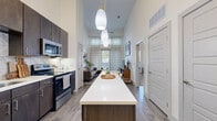 Located at 9330 Lookout Point in Dallas, TX, this two-bedroom, one-bathroom residence delivers a striking first impression with its dramatically high ceilings that amplify the sense of space throughout the open-concept main living area. The kitchen is a standout feature, outfitted with dark espresso cabinetry, stainless steel appliances, a chevron-patterned tile backsplash, white quartz countertops, and a generous elongated island with pendant lighting overhead — ideal for both meal prep and casual dining. A dedicated pantry provides additional storage convenience. The living room flows naturally from the kitchen, bathed in natural light from tall windows dressed with sheer patterned curtains, and is finished with wood-look plank flooring that runs throughout the main areas. The primary bedroom offers a comfortable, carpeted retreat with a large window framing outdoor views. Both bedrooms include closets. The bathroom features a dual-sink vanity with quartz countertops, dark cabinetry, wood-look flooring, and a full soaking tub with tiled surround. The cohesive design balances modern finishes with a warm, inviting ambiance throughout.