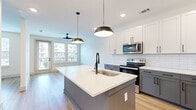 This contemporary single-floor residence showcases a thoughtfully designed open-concept layout with clean lines and modern finishes throughout. The kitchen is a standout feature, boasting two-tone cabinetry with crisp white upper cabinets and slate gray lower cabinets, accented by matte black hardware. A classic white subway tile backsplash complements the bright quartz countertops, while a spacious center island with an integrated sink and matte black faucet serves as the heart of the space. Stainless steel appliances, including a French door refrigerator, electric range, and over-the-range microwave, complete the kitchen. Stylish pendant lighting overhead adds warmth and definition to the area. Light wood-tone flooring flows seamlessly throughout the living areas, enhancing the bright and airy atmosphere created by large windows that frame views of the surrounding neighborhood. A ceiling fan in the living area adds both function and comfort. The bathroom is elegantly appointed with large-format gray tile surrounding the soaking tub and shower, a white vanity with quartz countertop, matte black fixtures, and direct access to a walk-in closet with built-in shelving. The overall ambiance is clean, modern, and move-in ready.